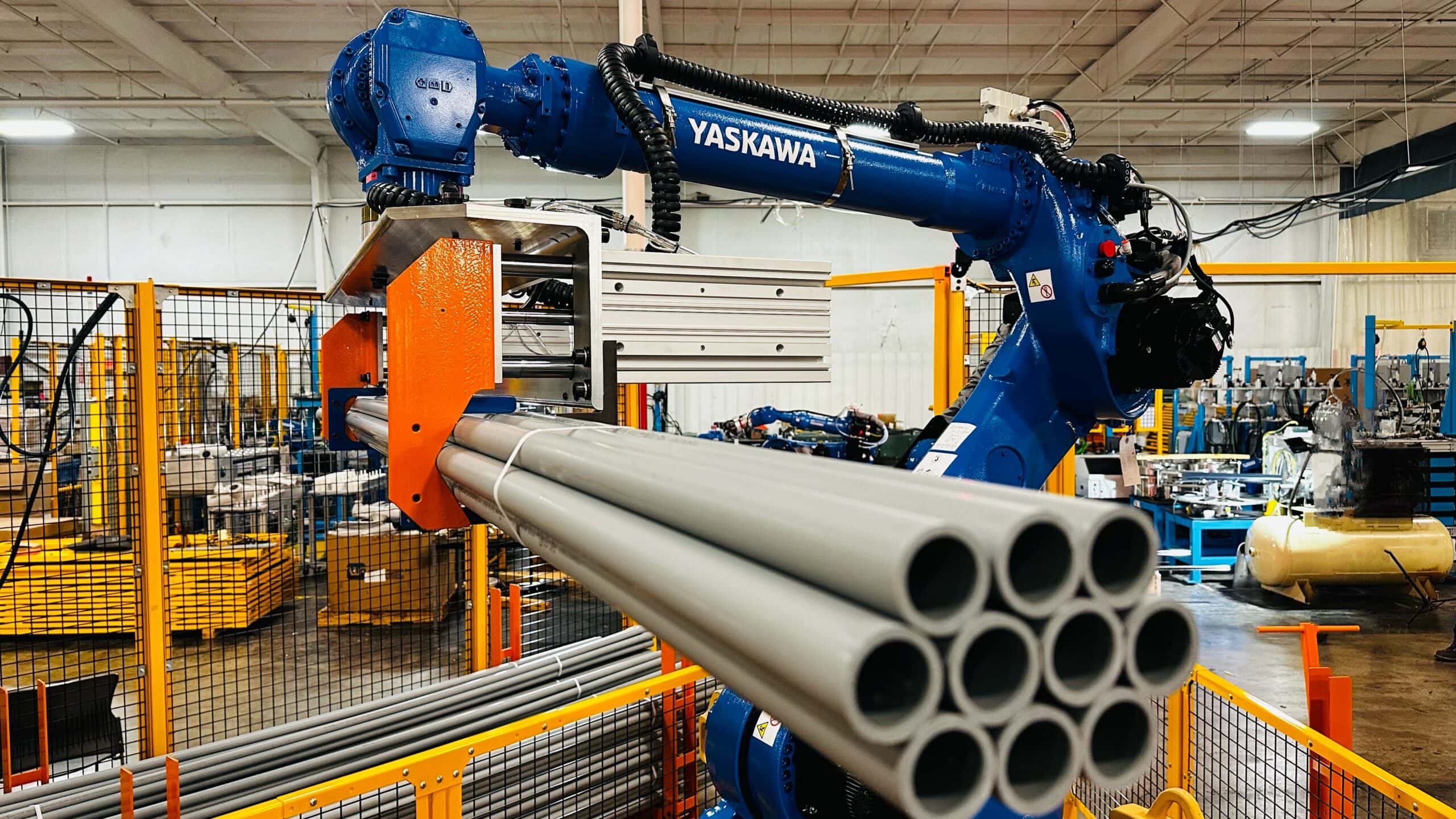 Yaskawa robotic arm handling gray pipes in an industrial setting, demonstrating automation in material handling and packaging processes.