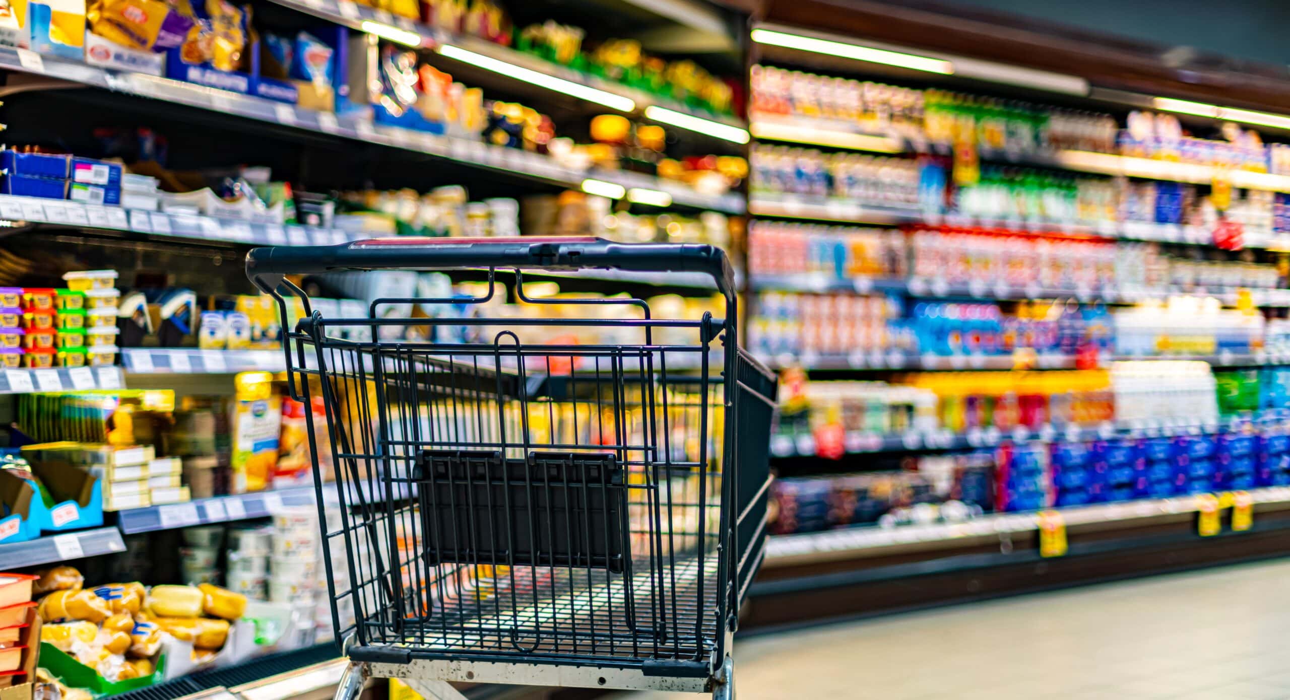 Shopping cart in grocery store aisle filled with consumer packaged goods, showcasing diverse products and packaging solutions relevant to automation systems for consumer goods production.